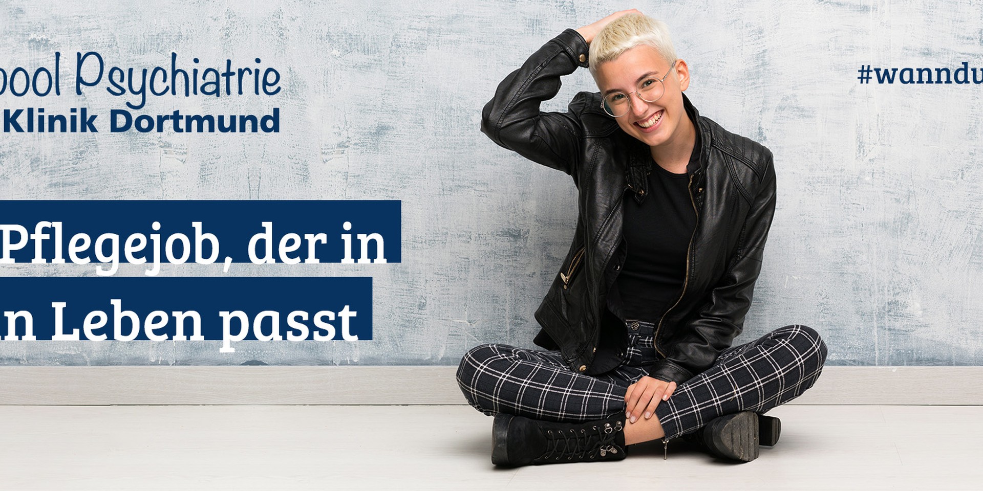Eine junge Frau sitzt im Schneidersitz vor einer Wand und hält die rechte Hand hinter ihrem Kopf